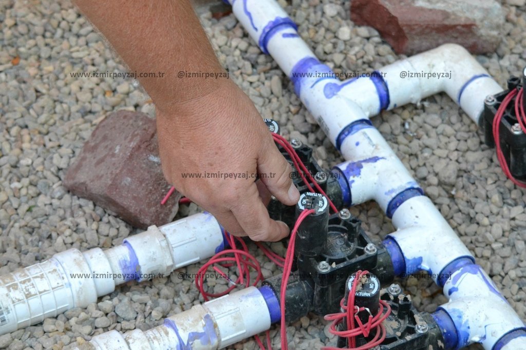 Bu fotoğrafta, bir bahçe sulama sisteminin PVC boruları ve vanaları görülmektedir. Bir el, vanalarda Bu fotoğrafta, bir bahçe sulama sisteminin PVC boruları ve vanaları görülmektedir. Bir el, vanalardan birini ayarlarken, sistemin kurulumu veya bakımı